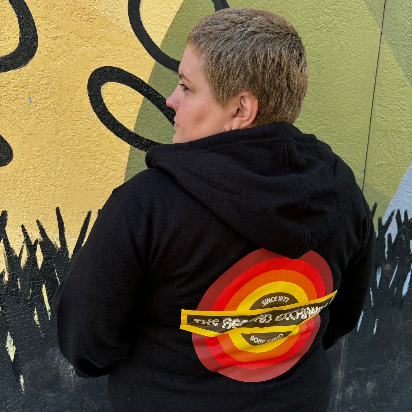 Model facing away showing Bright Retro Design of The Record Exchange's Newest Logo on Black Zip Up Hoodie.